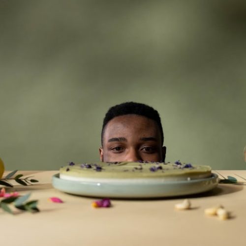 A man peeks over a table with cake, fruits, and flowers, creating a whimsical still life.