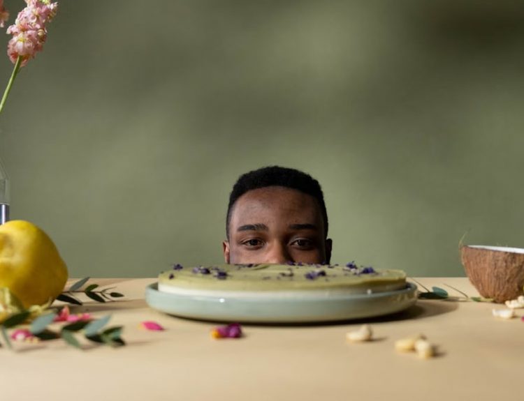 A man peeks over a table with cake, fruits, and flowers, creating a whimsical still life.