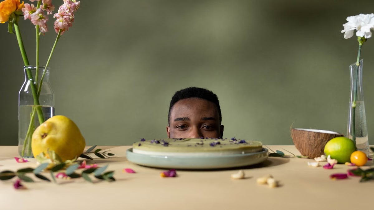 A man peeks over a table with cake, fruits, and flowers, creating a whimsical still life.