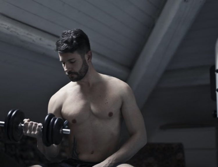 From below of muscular shirtless athletic male sitting on bench and doing bicep curls with dumbbell during intense training in dark studio
