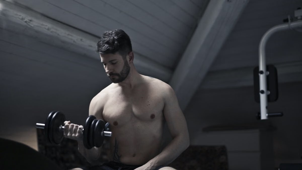 From below of muscular shirtless athletic male sitting on bench and doing bicep curls with dumbbell during intense training in dark studio